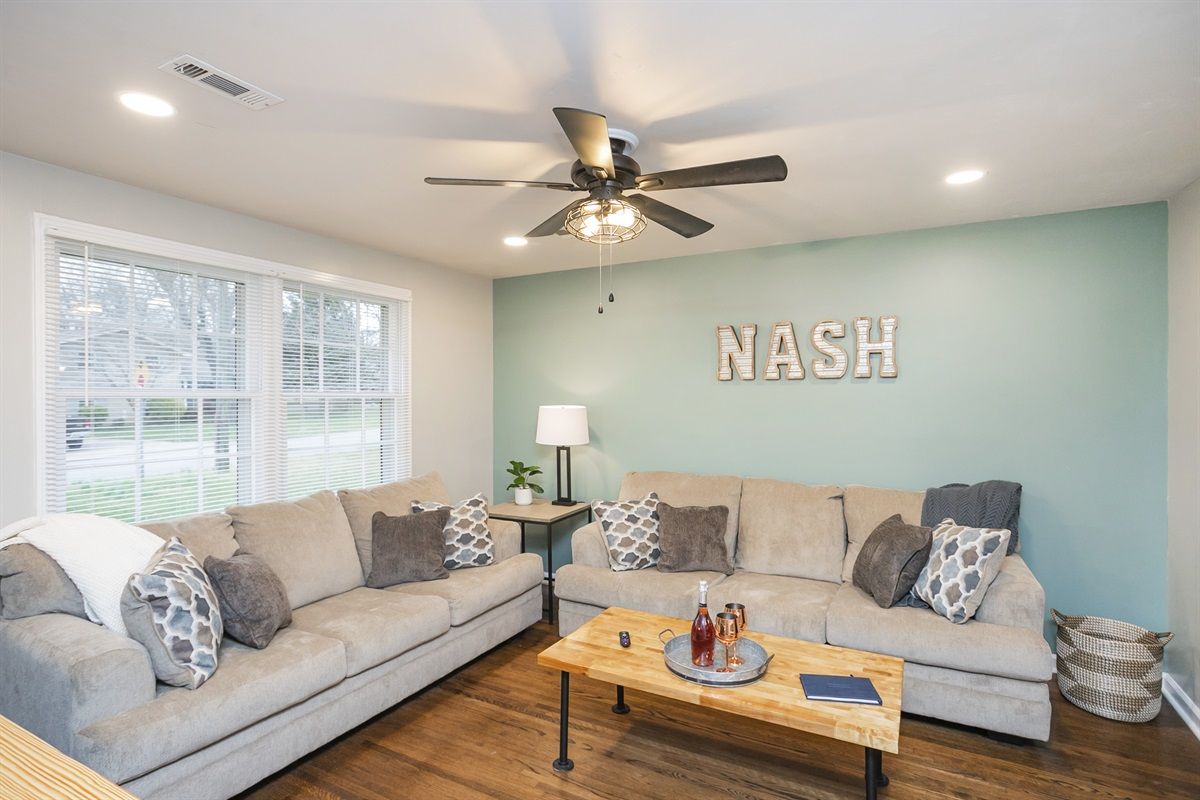 Bright, cozy modern living room with two beige sofas, patterned throw pillows, teal accent wall featuring 'NASH' letters, wooden coffee table, ceiling fan, hardwood floors and large front window.