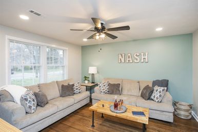 Bright, cozy modern living room with two beige sofas, patterned throw pillows, teal accent wall featuring 'NASH' letters, wooden coffee table, ceiling fan, hardwood floors and large front window.