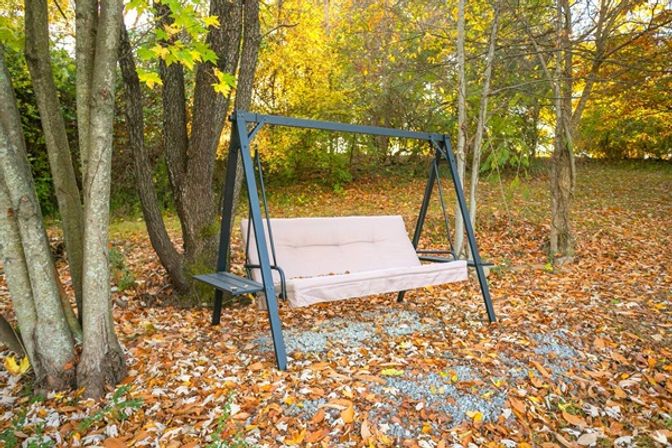 Cream-cushioned metal garden swing framed by trees in a backyard carpeted with orange and yellow autumn leaves