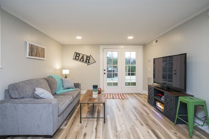 Bright cozy living room with gray sofa and turquoise throw, rustic wooden coffee table, light wood floors, French doors to backyard, 'BAR' arrow wall sign and flat-screen TV.
