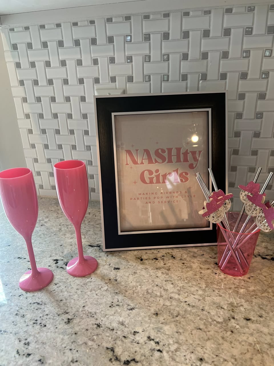 Kitchen countertop vignette with white basketweave tile backsplash, two bright pink plastic champagne flutes, a black-framed sign reading "NASHty Girls", and a pink cup holding glittery party stirrers.