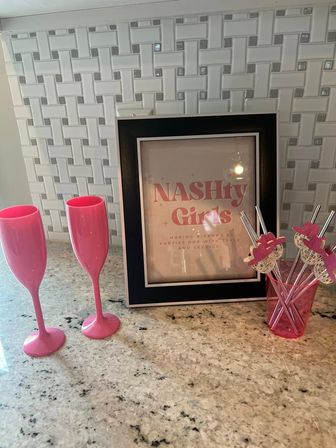 Kitchen countertop vignette with white basketweave tile backsplash, two bright pink plastic champagne flutes, a black-framed sign reading "NASHty Girls", and a pink cup holding glittery party stirrers.