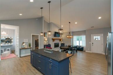 Bright open-concept kitchen and living area with a navy island and dark countertop, industrial pendant lights, vaulted ceiling, wood floors, dining table, fireplace and front entry