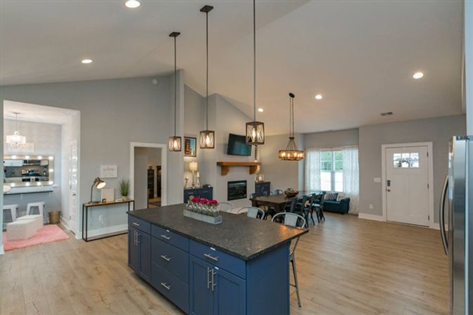 Bright open-concept kitchen and living area with a navy island and dark countertop, industrial pendant lights, vaulted ceiling, wood floors, dining table, fireplace and front entry