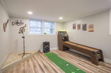 Finished basement game room with wooden shuffleboard, indoor putting green and clubs, small music stage with mic stand and speaker, light gray walls and wood-look flooring.