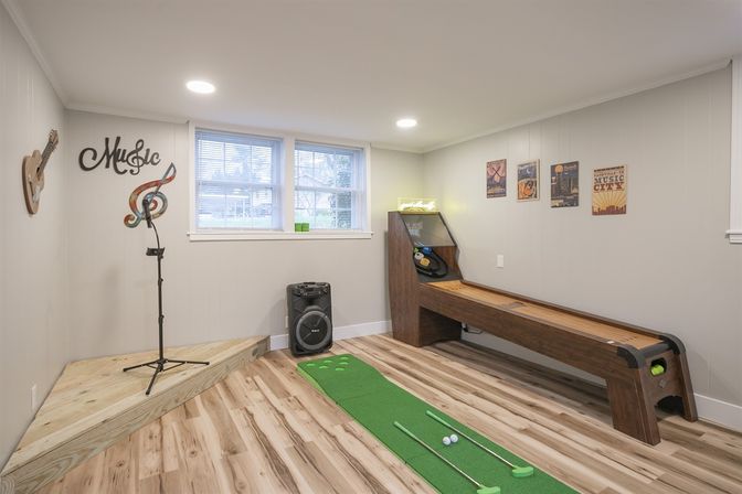Finished basement game room with wooden shuffleboard, indoor putting green and clubs, small music stage with mic stand and speaker, light gray walls and wood-look flooring.