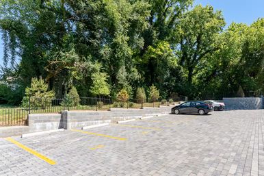 Sunlit suburban parking lot with gray interlocking pavers and yellow parking lines, a few parked cars, low concrete retaining walls and black metal fence bordering lush green trees under a clear blue sky.