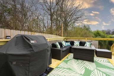 Cozy backyard wooden deck patio with covered gas grill, dark wicker sectional sofa with tropical-print pillows, rectangular fire-pit coffee table and green leaf-pattern outdoor rug overlooking a wooded yard and cloudy sky.