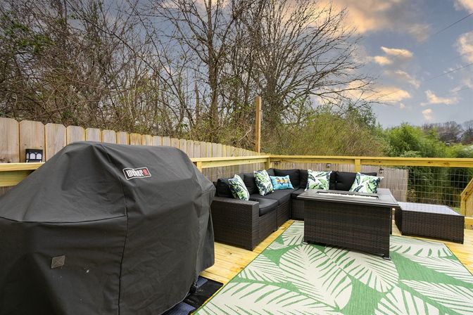 Cozy backyard wooden deck patio with covered gas grill, dark wicker sectional sofa with tropical-print pillows, rectangular fire-pit coffee table and green leaf-pattern outdoor rug overlooking a wooded yard and cloudy sky.