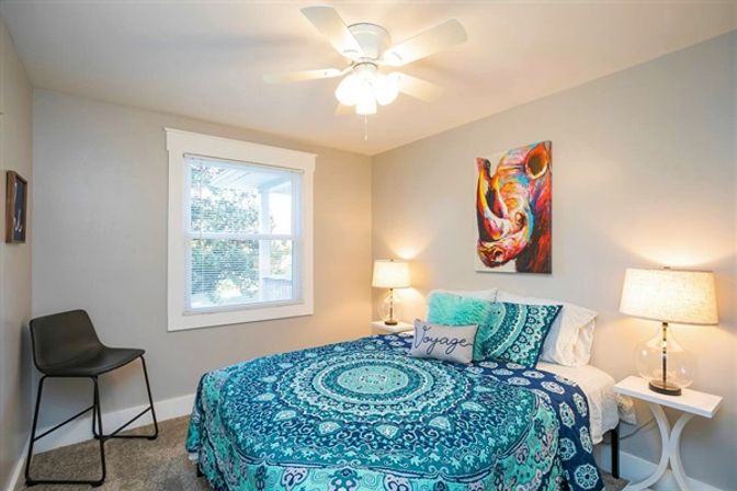 Sunny cozy bedroom with teal mandala bedspread, decorative pillows, vibrant rhino wall art, two bedside lamps on white nightstands, ceiling fan and a window with white trim.
