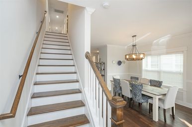 Sunlit modern home interior with a wooden staircase and white risers leading upstairs, adjacent dining area with a rectangular table, mixed patterned and upholstered chairs, a rustic chandelier, and dark hardwood floors.