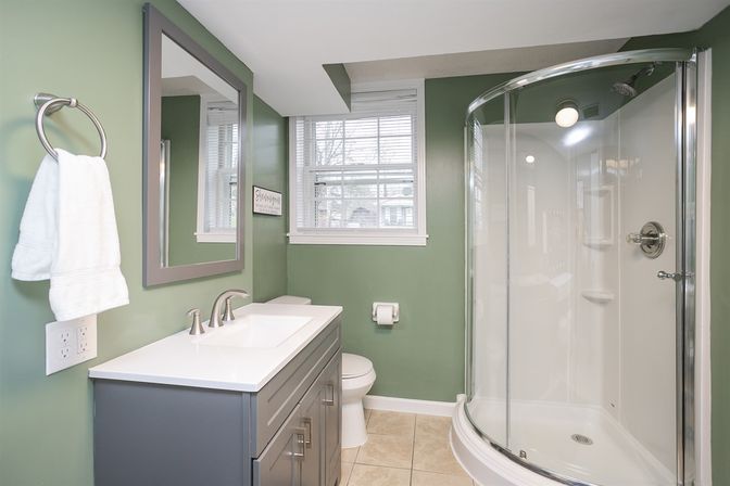 Bright green bathroom with curved glass corner shower, gray vanity with white countertop and sink, toilet, window and beige tile floor.