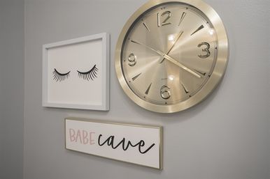 Modern gray wall gallery with a large gold round clock, framed eyelash illustration, and a rectangular 'Babe cave' sign.