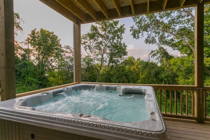 Bubbling hot tub on a covered wooden deck overlooking lush green trees and a pastel sunset sky