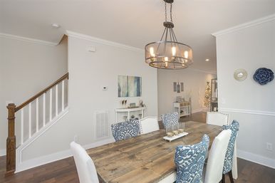 Cheerful modern dining room with a rustic reclaimed-wood table, blue patterned and white upholstered chairs, round metal chandelier, hardwood floors, white walls, and a nearby staircase—open, stylish interior.