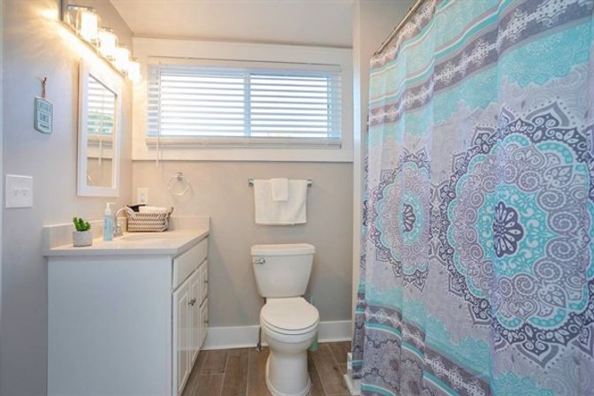 Bright modern bathroom with white vanity and sink, toilet, wood-look tile floor, and teal-gray mandala shower curtain under a window with blinds