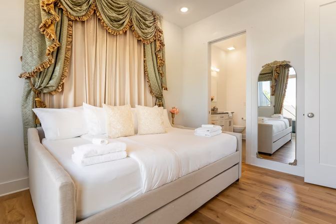 Bright guest bedroom with white linens and stacked towels, ornate green-gold canopy drapes, full-length mirror and ensuite bathroom on warm hardwood floors.
