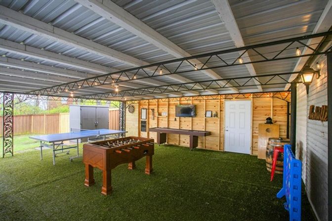 Covered backyard game room with metal roof and artificial turf, featuring a wooden foosball table, ping-pong table, long shuffleboard, wall-mounted TV and dartboard against pine-paneled wall, string lights and fenced yard