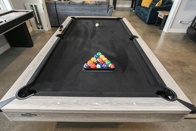 Modern indoor game room pool table with black felt and gray wood frame, colorful racked billiard balls at center and cue ball near the far end, concrete floor and lounge seating in the background.