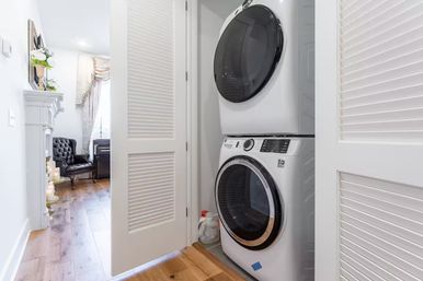 Bright home laundry nook with space-saving stacked white front-load washer and dryer in a louvered-door closet, hardwood floors and a peek into a cozy living room with tufted armchair and fireplace.