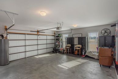Bright, organized two-car suburban garage with concrete floor, closed sectional garage door, wall cabinets and shelving, small workbench with two stools, lawn mower and stored tools, sunlight streaming through windows.