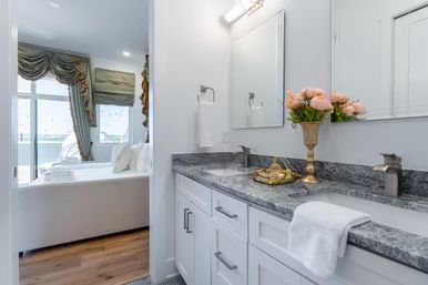Bright modern ensuite bathroom with double sinks, gray granite countertop and white shaker cabinets, a gold vase of peach flowers and decorative tray, towel draped over the counter, view into adjacent bedroom with draped windows and balcony.