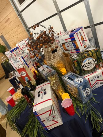 Party table in a garage with stacked beer cases (Michelob Ultra, Busch Light), wine bottles, red plastic cups, a woven vase filled with berry branches and evergreen table accents.