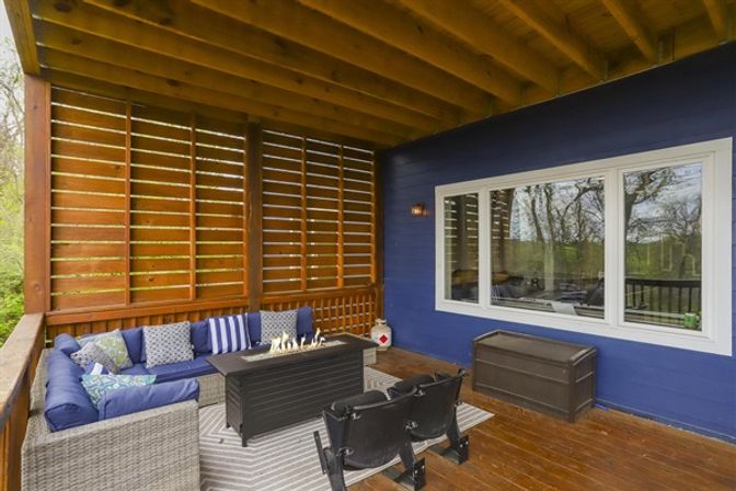 Cozy covered backyard deck with navy siding, wicker L-shaped sectional with blue cushions, rectangular gas fire-pit table, wooden privacy slats, two black seats and outdoor storage chest