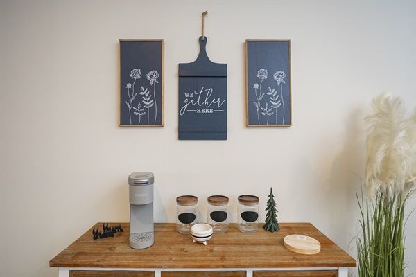 Modern farmhouse home beverage station on a wooden sideboard with a countertop beverage maker, three labeled glass jars with wooden lids, a small decorative tree and round wooden coaster, set beneath navy botanical wall art and tall pampas grass.