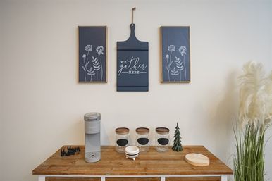 Modern farmhouse home beverage station on a wooden sideboard with a countertop beverage maker, three labeled glass jars with wooden lids, a small decorative tree and round wooden coaster, set beneath navy botanical wall art and tall pampas grass.
