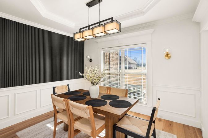 Modern bright dining room with wooden table and woven chairs, black round placemats and white vase of branches, vertical black accent wall, rectangular pendant chandelier, large window with blinds, white wainscoting and hardwood floors