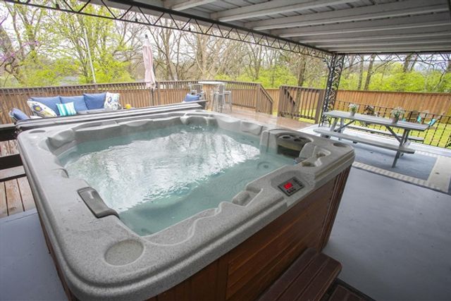 Covered backyard wooden deck with a large bubbling hot tub, cushioned outdoor sofa, picnic table and bar stools overlooking a leafy treed yard.