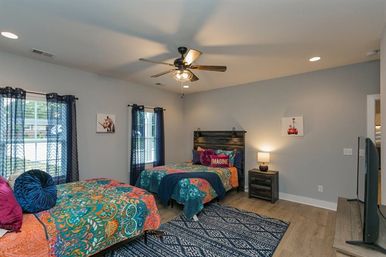 Bright modern shared bedroom with two twin beds in colorful bohemian bedding, navy curtains over windows, wood floors, patterned area rug, ceiling fan, nightstand with lamp and decorative pillows including one that reads 'IMAGINE'.