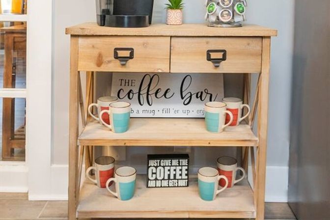 Home coffee bar on a light wooden console table with two drawers, single-serve coffee maker and pod carousel on top, decorative coffee signs, and colorful turquoise and coral ceramic mugs neatly arranged on open shelves.