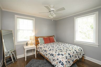Bright cozy bedroom with gray walls, hardwood floors, wooden bed dressed in floral duvet and colorful throw pillows, white bedside table with lamp, leaning full-length mirror, two large windows and ceiling fan.