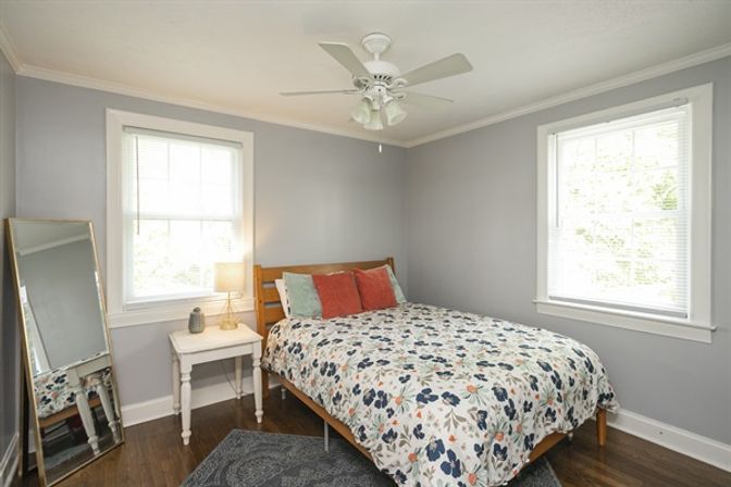Bright cozy bedroom with gray walls, hardwood floors, wooden bed dressed in floral duvet and colorful throw pillows, white bedside table with lamp, leaning full-length mirror, two large windows and ceiling fan.