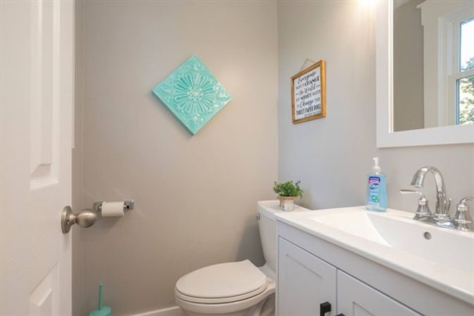 Small residential bathroom with white vanity and chrome faucet, toilet, neutral gray walls, teal decorative wall tile and framed sign, potted succulent and bottle of hand soap on the sink