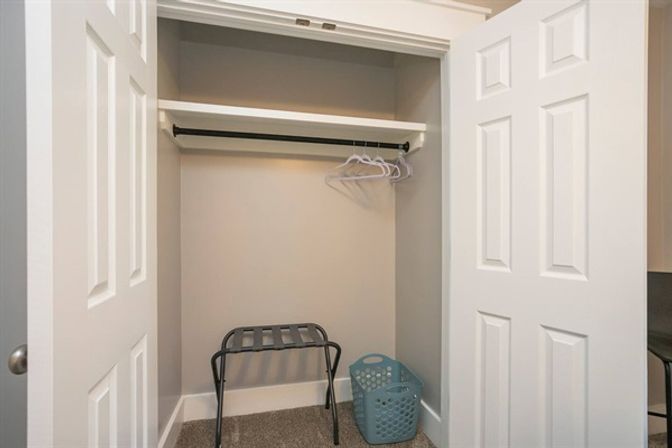 Open bedroom closet in home with white doors, shelf and black hanging rod holding empty plastic hangers, carpeted floor with a metal luggage rack and teal laundry basket.