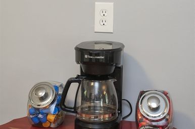 Countertop coffee station with a black drip coffee maker and glass carafe, flanked by two glass jars—one filled with colorful single‑serve pods and one with coffee beans—set against a gray wall with a white outlet.