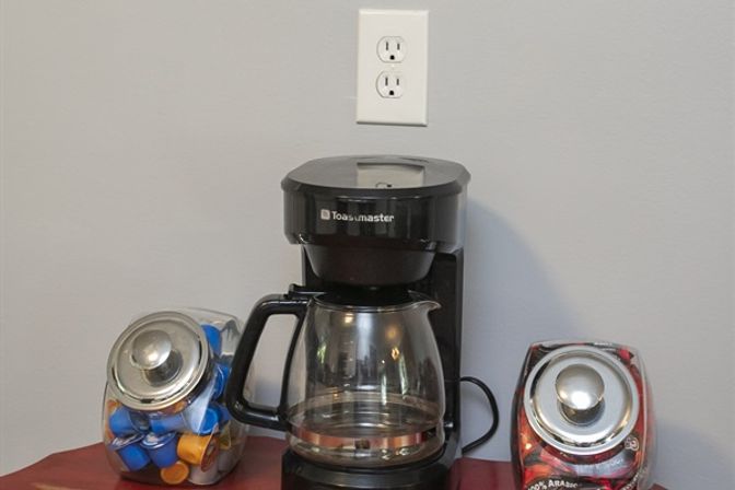 Countertop coffee station with a black drip coffee maker and glass carafe, flanked by two glass jars—one filled with colorful single‑serve pods and one with coffee beans—set against a gray wall with a white outlet.
