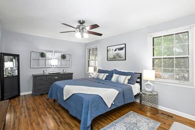 Cozy light-filled master bedroom with hardwood floors, blue quilted bedding on a king-size bed, ceiling fan, gray dresser, bedside lamps and large windows