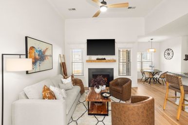 Bright open-plan living room interior with white walls and wood floors, wall-mounted TV above a fireplace, light-gray sofa with throw pillows, two brown leather club chairs, wooden coffee table, abstract guitar wall art, and a dining nook in the background.