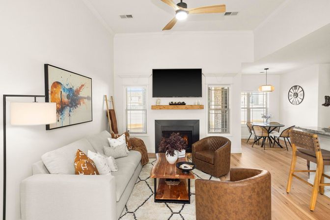 Bright open-plan living room interior with white walls and wood floors, wall-mounted TV above a fireplace, light-gray sofa with throw pillows, two brown leather club chairs, wooden coffee table, abstract guitar wall art, and a dining nook in the background.