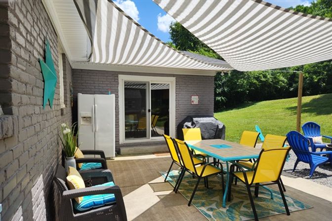 Cheerful backyard patio with striped shade sails, turquoise dining table surrounded by six yellow chairs on an outdoor rug, wicker lounge chairs with blue cushions, sliding glass door, fridge and covered grill next to a green lawn.