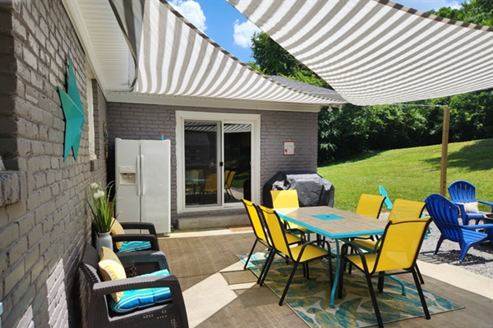 Cheerful backyard patio with striped shade sails, turquoise dining table surrounded by six yellow chairs on an outdoor rug, wicker lounge chairs with blue cushions, sliding glass door, fridge and covered grill next to a green lawn.