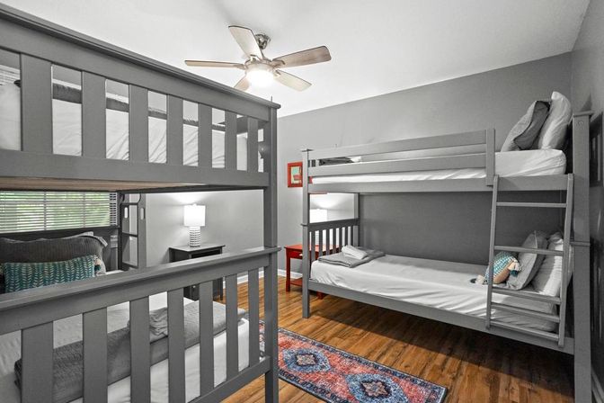 Cozy modern bedroom with two gray twin bunk beds, hardwood floors, ceiling fan, colorful runner rug, red nightstand and lamp — bright kids' or guest room.