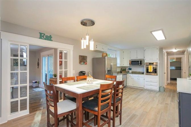 Bright open-concept kitchen and dining area with a wooden high-top table and six chairs, white cabinets, stainless steel refrigerator and built-in oven, pendant lights, wood-look tile floor, and glass-paneled doorway