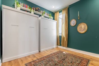 Cozy modern bedroom with teal accent wall, two white Murphy beds, hardwood floors and patterned area rug, gold window curtains, floating shelf of books and trailing plants, and round vintage wall plaques.