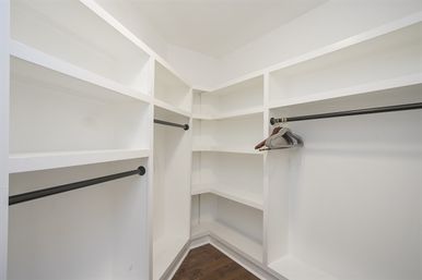 Bright walk-in closet with white built-in shelving and corner cubbies, black metal clothing rods with a few wooden hangers, and dark hardwood flooring.