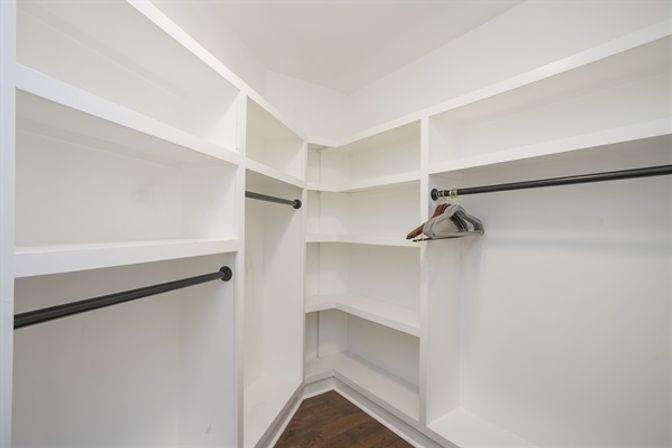 Bright walk-in closet with white built-in shelving and corner cubbies, black metal clothing rods with a few wooden hangers, and dark hardwood flooring.